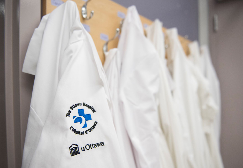 White lab coats hanging on hooks, one showing Ottawa Hospital and uOttawa logos.