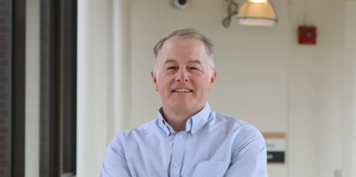 A smiling man stands in a hallway with his arms crossed