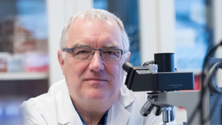 A man sits in a lab and smiles at the camera with a microscope to the right of his face