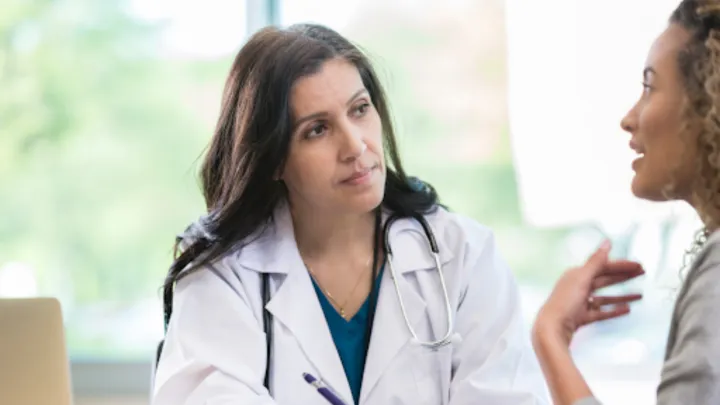 Two woman (one a doctor wearing a lab coat) sit at table and speak