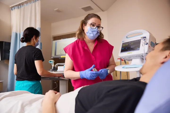 One nurse is standing at a computer while another nurse is in front of a patient and speaking with him