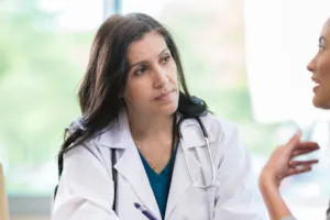 Two woman (one a doctor wearing a lab coat) sit at table and speak