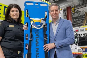 A woman and a man stand in front of two ambulances and hold a blue backboard