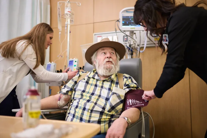 An older person in a plaid shirt and hat sits in a medical chair while two healthcare workers take blood pressure and adjust an IV.