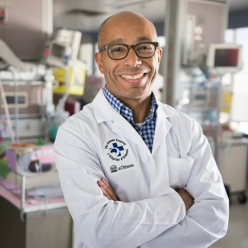 A man wearing a lab coat and glasses stands with his arms crossed in a lab