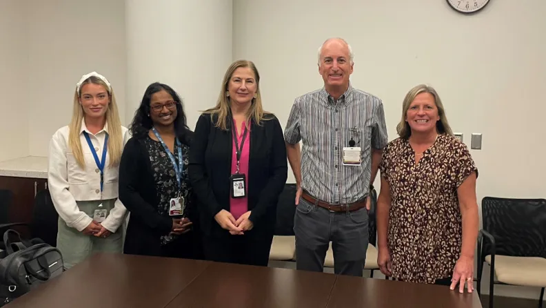 Five people stand together in an meeting room and smile