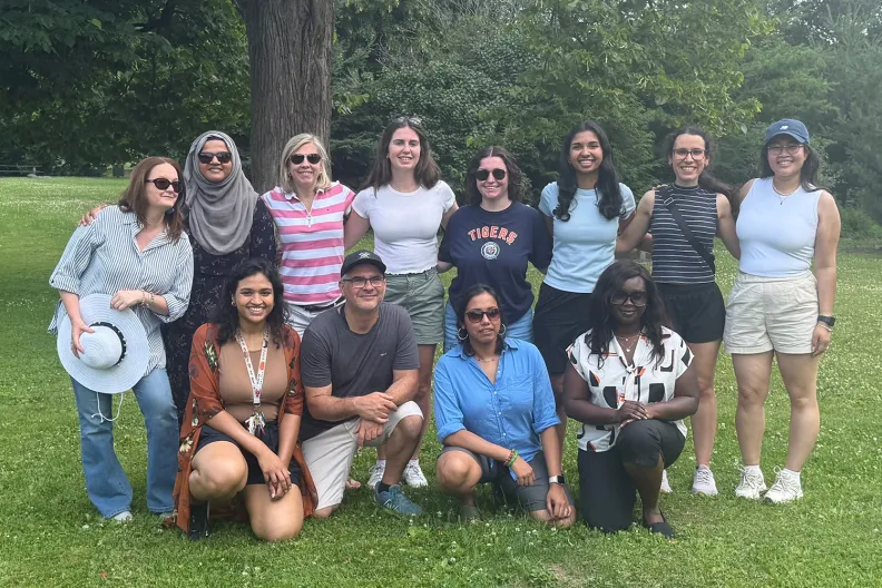 Twelve people posing for the camera with grass and trees in the background.